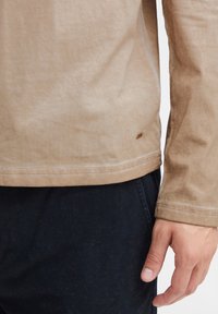 Pitkähiha beige paita, jossa on teksturoitu pinta ja pieni logo-etiketti helman lähellä. Yhdistetty tummaan housuun, näkyvillä käsi.