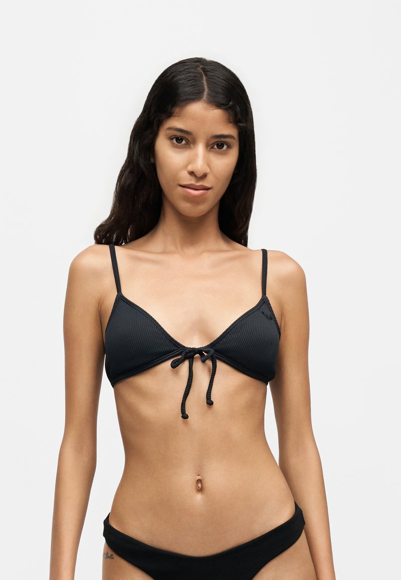 Femme aux longs cheveux foncés portant un bikini noir à fines bretelles et un nouage devant, debout sur un fond clair uni.