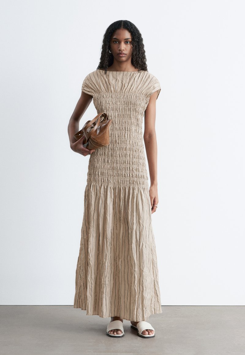 Une femme se tient debout, vêtue d'une robe longue beige texturée et de sandales blanches sans lacets, tenant un sac à main en cuir marron sur un fond blanc uni.