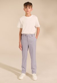 Băiat purtând un tricou alb, pantaloni gri deschis și adidași albi, stând cu o mână în buzunar pe un fundal bej.