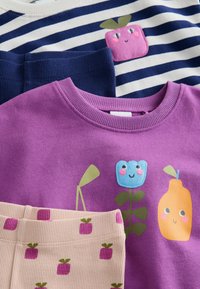 Drei farbenfrohe Kleidungsstücke: ein lila Sweatshirt mit floralen Designs, ein gestreiftes Oberteil in Marineblau und Weiß mit einem pinkfarbenen Quadratmotiv und beige Ripp-Pants mit kleinen pinken Mustern.