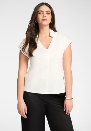 Vrouw met lang bruin haar, gekleed in een witte V-hals korte mouwen blouse en zwarte broek, staand tegen een effen achtergrond.