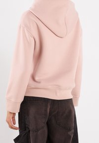 Sweat à capuche rose clair avec poignets et ourlet côtelés, doté d'une texture douce. Il est porté avec un pantalon cargo marron foncé.