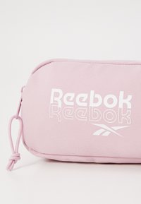 Pochette en tissu texturé rose mettant en avant le logo « Reebok » en blanc et une fermeture éclair avec une tirette nouée.