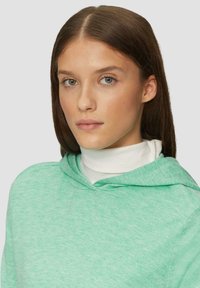 Mintgrüner Kapuzenpullover aus weichem, strukturiertem Stoff mit lässiger Passform. Darüber getragen, zeigt er einen weißen Rollkragen, der ein schlichtes Design aufweist.