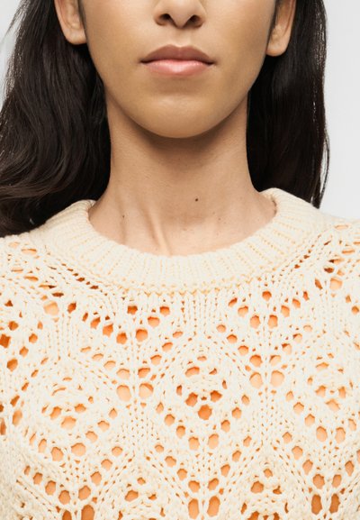 Gros plan d'une personne à la peau de ton moyen portant un pull en maille clair décontracté par-dessus une chemise pêche, ne montrant que le cou et le bas du visage.