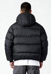 Zwarte pufferjas met capuchon, voorzien van horizontale stiksels en elastische manchetten. Zacht, glanzend materiaal en een ontwerp met een ontspannen pasvorm.