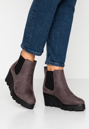 Dames grå ruskind kilehæl støvler med sorte elastiske sidelommer båret med opkantsede blå jeans på en hvid baggrund.