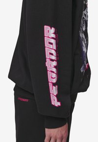 Manche de sweat-shirt noire avec un texte stylisé en rose vif et blanc indiquant "PETORR" et des caractères japonais plus petits à côté.