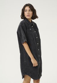 Vestido camista de mezclilla negra con mangas cortas, cierre de botones en la parte frontal y dos bolsillos en el pecho. Corte ligeramente holgado y dobladillo curvado para un estilo añadido.