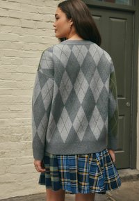 Kvinde står udendørs med ryggen til, iført en grå argyle sweater og en blåternet plisseret nederdel, nær en grøn dør og en sandfarvet murstensvæg.