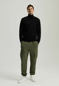 Czarny sweter z golfem połączony z zielonymi spodniami cargo z dużymi bocznymi kieszeniami; dopełniony białymi sneakerami. Prosty, dopasowany design.