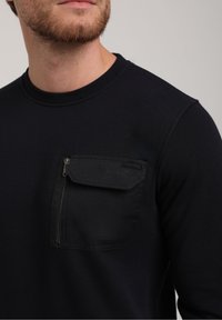 Navy blue sweatshirt with a crew neckline, featuring a zippered chest pocket made of a textured fabric. Smooth texture overall.