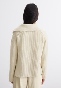 Pull en maille côtelée de couleur crème avec un col replié et des épaules tombantes, présentant une coupe décontractée et une texture douce.