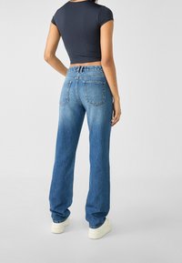 Žena v přímých modrých džínách, černém crop topu a bílých teniskách, stojící zády k fotoaparátu na neutrálním pozadí.