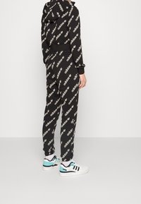Černý teplákový komplet s celoplošným vzorem loga Adidas v bílé barvě, zahrnující mikinu s kapucí a odpovídající zúžené kalhoty, doplněný o bílé tenisky.