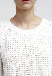 Bílý pletený svetr s kulatým výstřihem a otevřenou texturou, který představuje volný a prodyšný design.