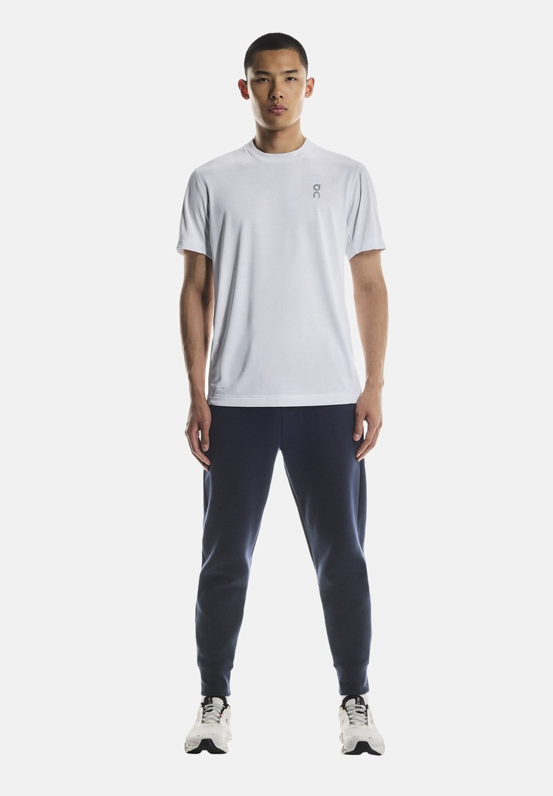 Weißes kurzärmeliges Sport-T-Shirt mit kleinem Logo auf der linken Brust, kombiniert mit marineblauen Jogginghosen und weißen Sportschuhen.