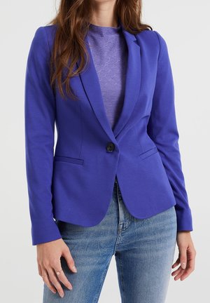 Vrouw draagt een nauwsluitende koningsblauwe blazer over een gestructureerde paarse top, gecombineerd met lichtblauwe spijkerbroek, tegen een effen achtergrond.