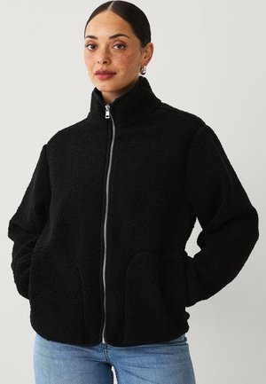 Schwarze Fleecejacke mit hohem Kragen, vollständig durchgehendem Reißverschluss und seitlichen Taschen. Die Jacke verfügt über einen weichen, strukturierten Stoff und eine lässige Passform.