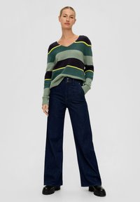 Mehrfarbiger, grün-, marineblau- und gelbgestreifter Strickpullover mit V-Ausschnitt; weite, dunkelblaue Jeans; schwarze klobige Stiefel.