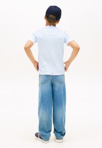 Αγόρι που στέκεται με τα χέρια στη μέση, φοράει μπλε σκούφο, ανοιχτό μπλε μπλουζάκι, φαρδιά μπλε τζιν και αθλητικά παπούτσια, μπροστά από λευκό φόντο.