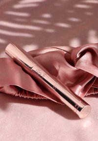Un tube de mascara en métal rose brillant repose sur un tissu satin léger rose pâle, affichant un design élégant et une texture lisse.