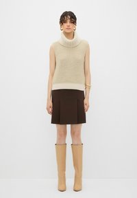 Maglione smanicato beige a collo alto, sovrapposto a una mini gonna marrone, abbinato a stivali alti color cammello e orecchini a cerchio dorati.