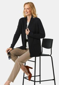 Schwarze Strickjacke mit Schalkragen, gerippten Bündchen und Vordertasche. Kombiniert mit beigen Hosen und braunen Wildlederabsätzen, sitzend auf einem schwarzen Stuhl.