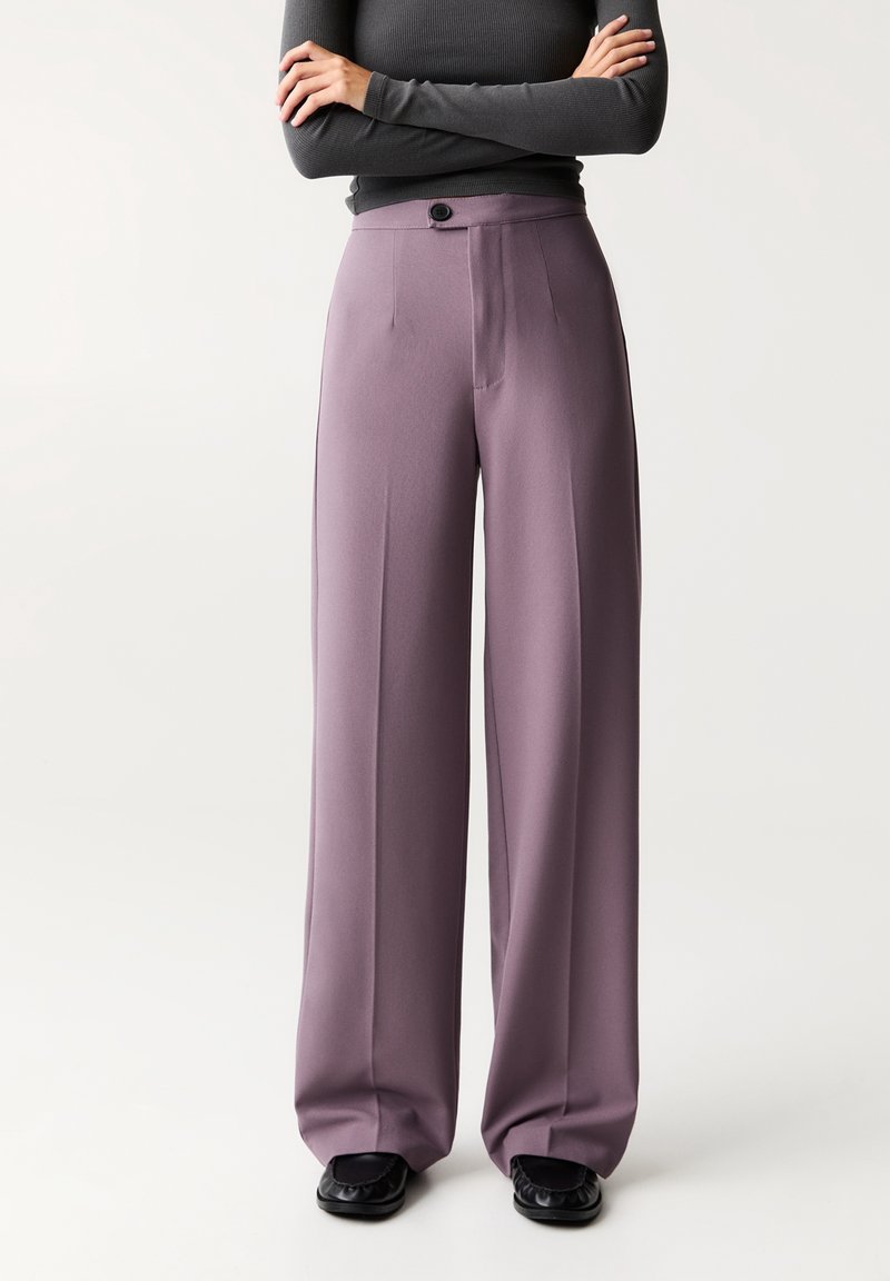 Personne portant un pantalon mauve taille haute à jambes larges, des mocassins noirs et un haut ajusté à manches longues gris foncé, les bras croisés.