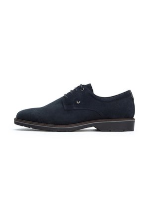 Zapato de vestir de ante azul marino con punta redondeada, diseño con cordones y suela fina marrón. Presenta un sutil logo en el lateral.