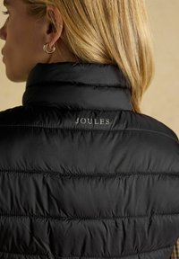 Fekete puffkabát magas gallérral, steppelt textúrával és látható varrással. A háttéren kontrasztos színű "JOULES" logó található.