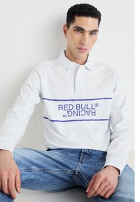 Balta gara piedurknē polo krekls ar zilu horizontālu joslu, uz kuras ir teksts "RED BULL RACING", komplektā ar ziliem džinsiem.