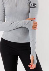 Maglietta grigia a maniche lunghe con coste testurizzate, collo con zip a metà e aperture per il pollice. Presenta una patch con il logo sulla manica sinistra e un design aderente.