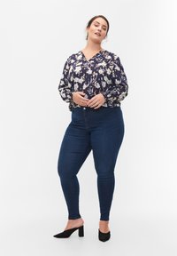 Blusa navy con estampado floral, escote en V y puños elásticos, combinada con jeans ajustados de un azul oscuro. La modelo lleva zapatos negros de tacón bloque.