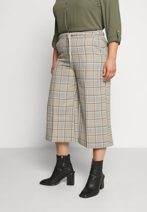 Vrouw draagt beige geruite wijde cropped broekspijpen, een olijfgroen shirt met lange mouwen en zwarte enkellaarsjes met hak, staand tegen een effen achtergrond.