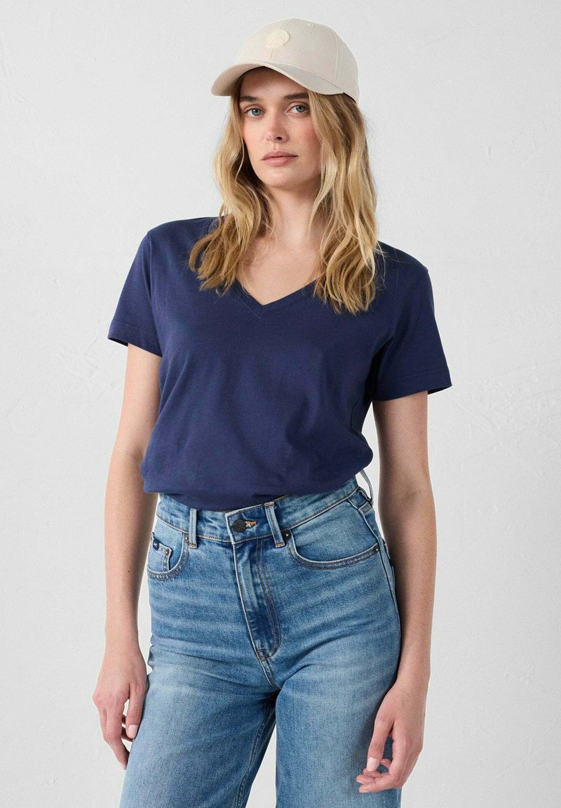 Mujer con cabello largo y rubio que lleva una gorra beige, una camiseta azul marino de cuello en V y unos pantalones vaqueros de tiro alto en azul claro, frente a un fondo blanco.