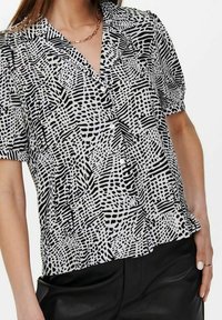 Blouse à manches courtes en tissu noir et blanc avec des motifs abstraits. Dotée d'un col en V et d'une coupe ample, associée à un pantalon noir.
