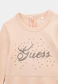 Persiku krāsas krekls ar apaļu kakla izgriezumu, kas rotāts ar "Guess" sudraba pērlītēm un izkaisītiem sudraba uzlīmiem papildu detaļām. Aizmugurējā rāvējslēdzēja aizdare.