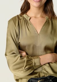 Woman wearing olive green textured blouse with gathered cuffs and stand collar, arms crossed over black belt and jeans.