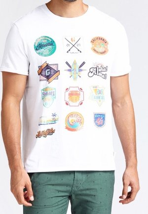 T-shirt blanc avec des logos vintage colorés, y compris Hawaii, Californie, planche de surf, Aloha et des thèmes estivaux, porté par un homme debout.