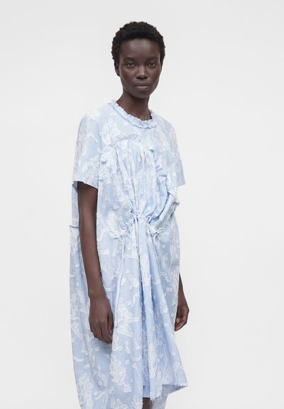 Γυναίκα με σκουρόχρωμο δέρμα που φοράει ανοιχτό μπλε φόρεμα με λευκά ανθισμένα μοτίβα και λεπτομέρεια κόμπου, στέκεται μπροστά σε απλό φόντο.