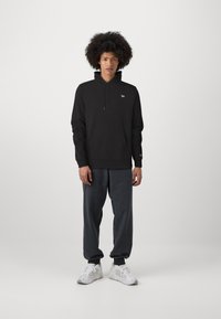 Felpa nera con cappuccio e tasca frontale, abbinata a pantaloni jogger grigi scuri. Sneakers bianche completano l'outfit. Presenti pochi loghi.