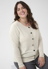 Cardigan de culoare crem, cu decolteu rotund, mâneci lungi și nasturi negri în față. Manșetele și tivul ribbate adaugă textură.