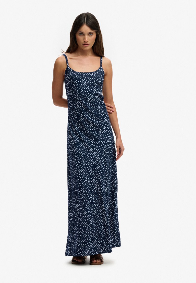 Kvinde iført en marineblå, ankel-længde kjole med hvide polkaprikkker og tynde skulderstropper, stående mod en ensfarvet baggrund.