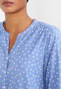 Blouse bleu clair avec un col en V boutonné, présentant des motifs floraux blancs et des détails plissés aux épaules. Texture de tissu douce.