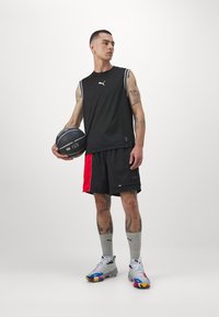 Μαύρο αμάνικο μπλουζάκι μπάσκετ με άσπρες ρίγες, συνδυασμένο με μαύρες σορτς που διαθέτουν ένα κόκκινο πάνελ. Γκρι αθλητικές κάλτσες και πολύχρωπα αθλητικά παπούτσια.