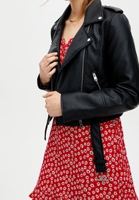 Veste de moto en faux cuir noir avec des fermetures éclair argentées et une ceinture bouclée, portée sur une robe florale rouge ornée de marguerites blanches.
