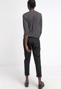 Frau, die mit dem Rücken zur Kamera steht, trägt eine schwarze Bluse mit Muster und drei Viertelärmeln, schwarze 7/8-Hosen und schwarze Schuhe.