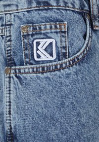 Denim jeans in light blue with a distressed texture. Features a white embroidered logo on the back pocket and orange stitching details.
